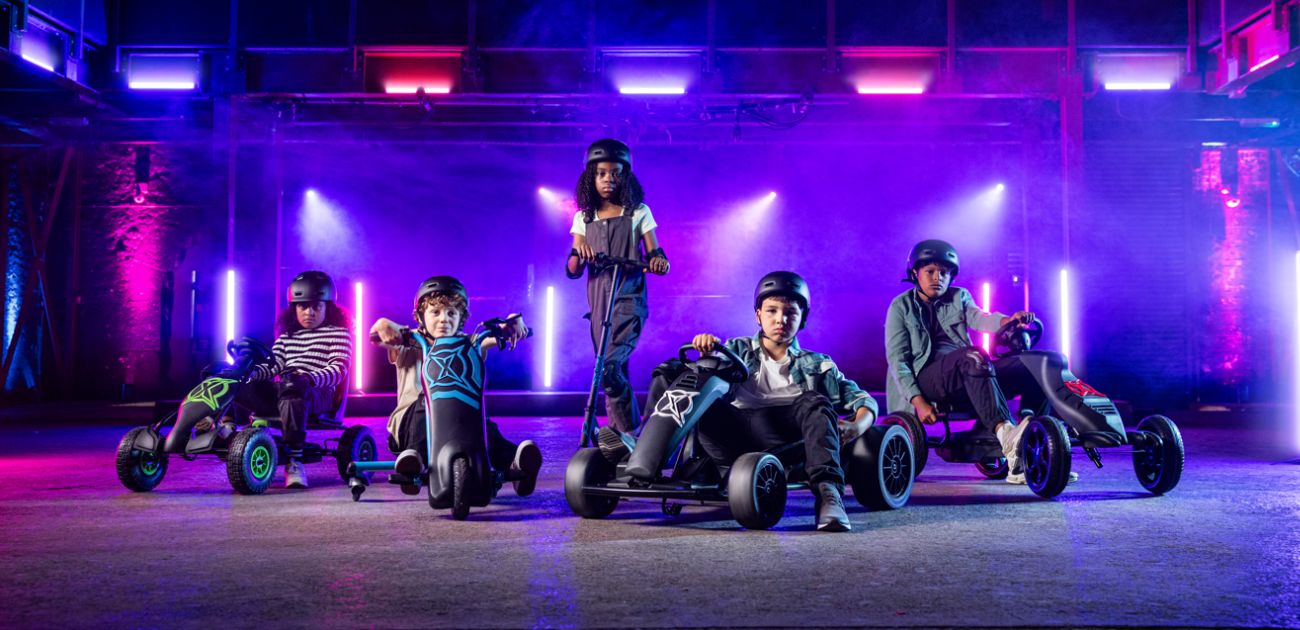 A photo of a group of children with go karts and scooters. They are in a warehouse lit with pink and purple lights