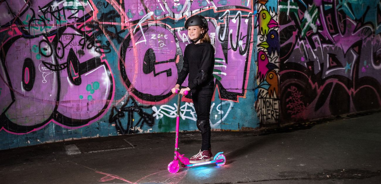 A photo of a girl riding a pink scooter with an blue LED deck. The background is pink and blue graffiti