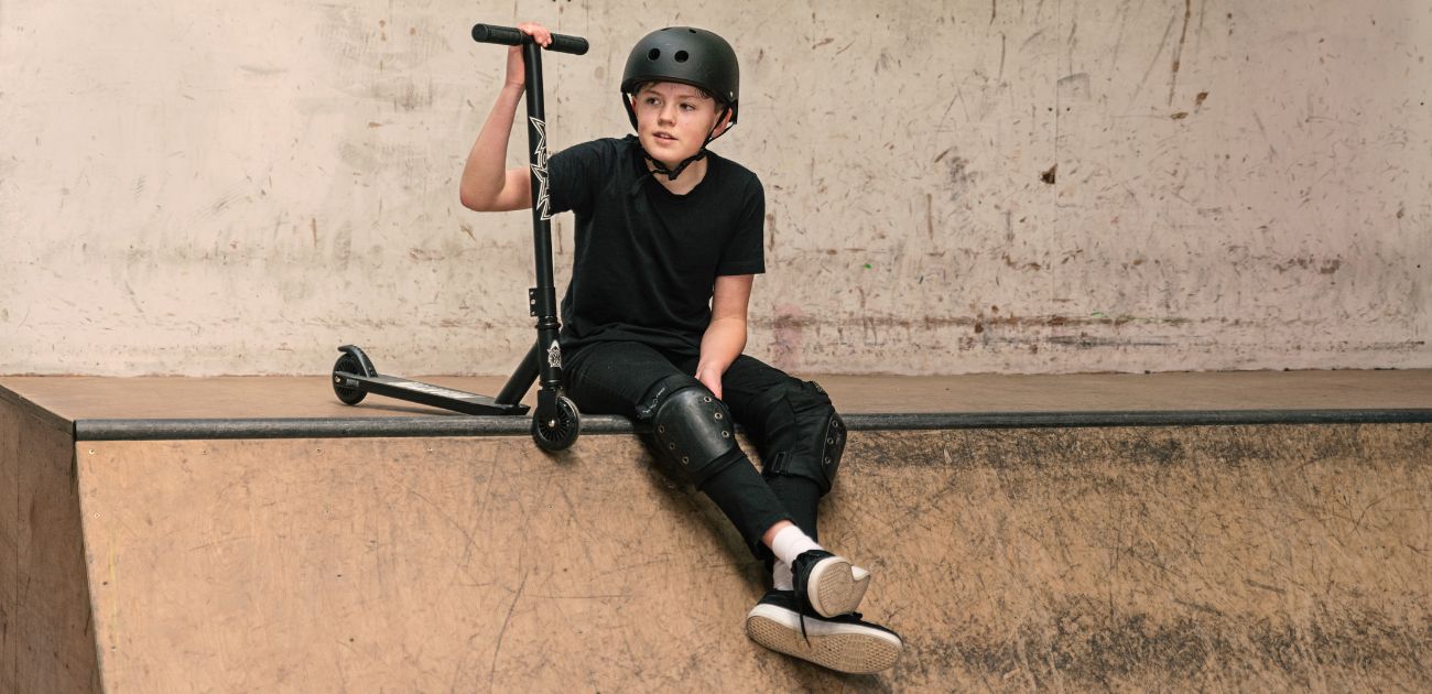 A photo of a boy with a black stunt scooter sitting on the edge of a skate ramp