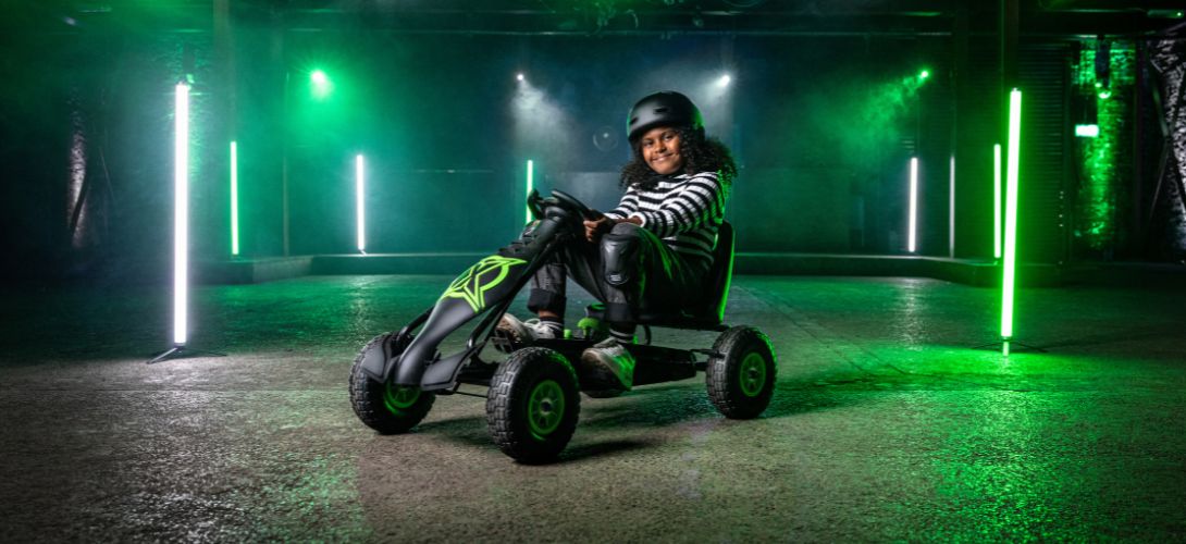 A photo of a girl riding a black and green go kart. She is in a dark warehouse which is lit with white and green lights