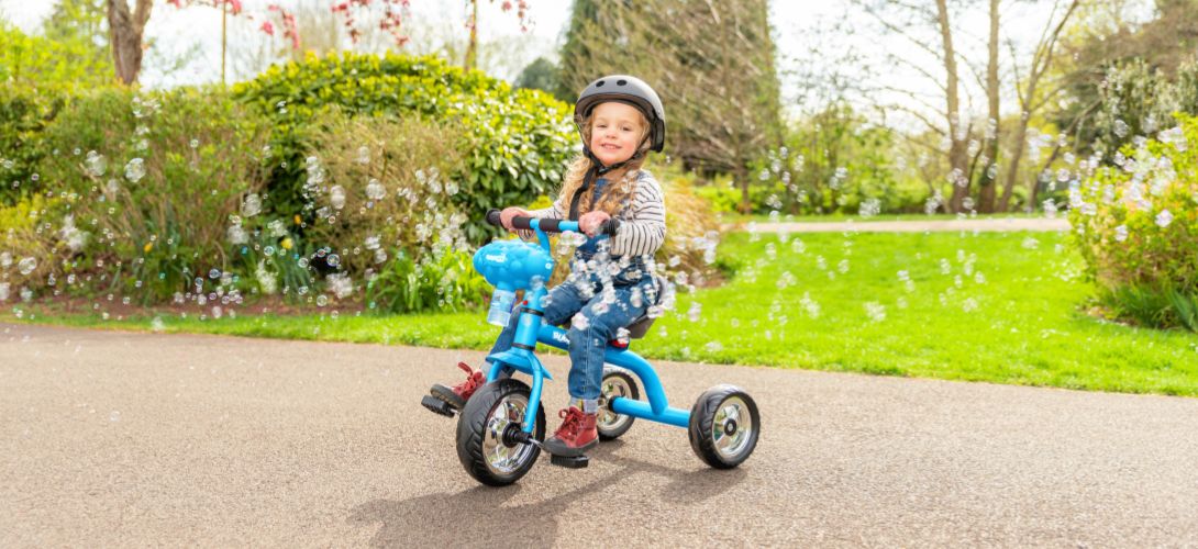 A photo of a girl riding a blue trike through a park with a bubble machine on the front