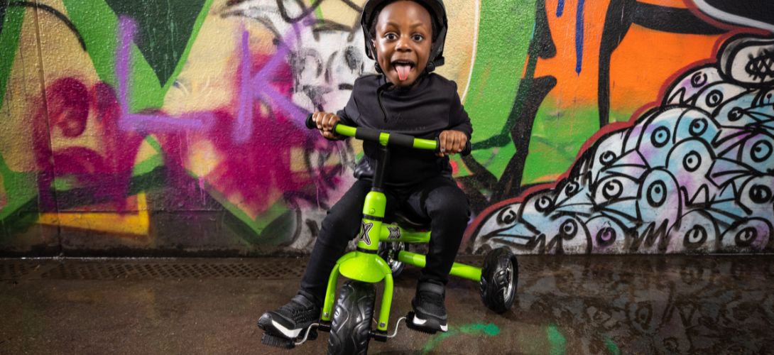 A photo of a young boy riding a green trike. He is in front of a graffiti background and sticking his tongue out