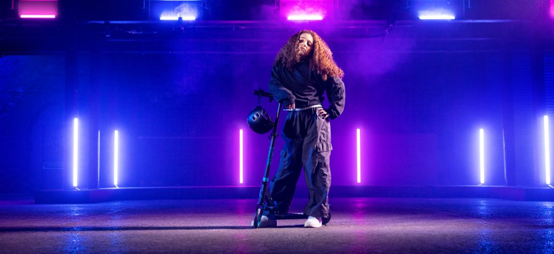 A photo of a girl in a warehouse which is lit by purple and pink lights. She is posed with a black electric scooter 