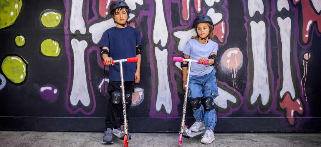 A photo of a boy and a girl holding scooters. They are stood against a dark graffiti background