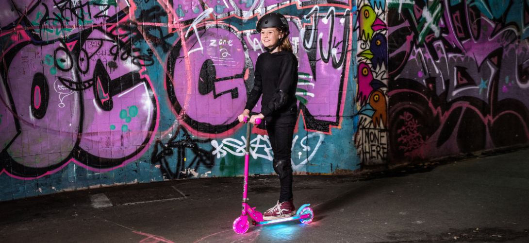 A photo of a girl riding a pink and blue scooter with an LED deck against a pink graffiti wall