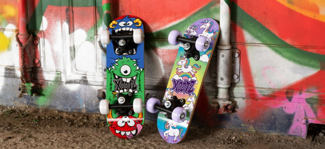 A photo of two mini skateboards against a graffiti background. One has a unicorn print deck, the other has a monster print